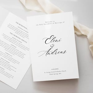 以下が含まれることがあります： 黒い文字が書かれた白いウェディングプログラム。1つは開いており、イベントの順序が表示され、もう1つは閉じており、「Eleni & Andreas」の名前が筆記体で表示されています。クリーム色のリボンが近くにあります。