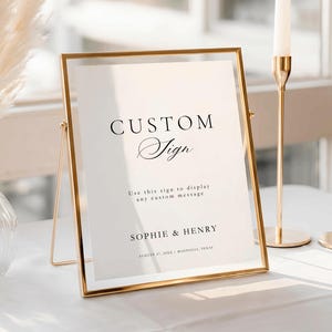 Può includere: Un cartello con cornice dorata con le parole "CUSTOM Sign" in elegante scrittura. Il cartello include il testo "Use this sign to display any custom message" e i nomi "SOPHIE & HENRY". Due candelabri dorati e un vaso sullo sfondo.