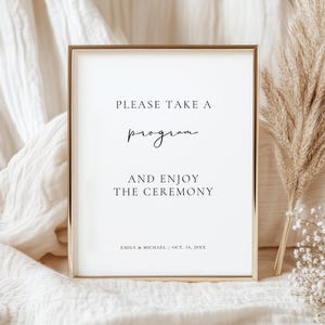 May include: A framed wedding ceremony sign with a gold border. The sign displays the text "PLEASE TAKE A program AND ENJOY THE CEREMONY" in black script. The names "EMILY & MICHAEL" and the date "OCT. 14, 20XX" are at the bottom.