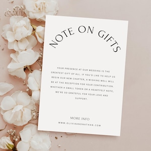 Puede incluir: Una tarjeta color crema con el texto "NOTE ON GIFTS" en escritura negra. La tarjeta incluye texto relacionado con bodas y el sitio web www.oliviaandnathan.com. Flores blancas y vegetación dispuestas alrededor de la tarjeta.