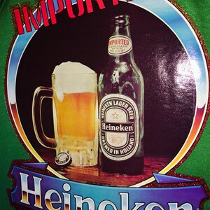 Pu&ograve; includere: Una maglietta verde con un design di glitter dorati che presenta un bicchiere di birra e una bottiglia di birra Heineken. Il testo "IMPORTED" &egrave; in lettere rosse e il testo "Heineken" &egrave; in lettere di glitter dorati.