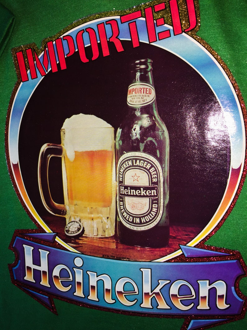 Pu&ograve; includere: Una maglietta verde con un design di glitter dorati che presenta un bicchiere di birra e una bottiglia di birra Heineken. Il testo "IMPORTED" &egrave; in lettere rosse e il testo "Heineken" &egrave; in lettere di glitter dorati.