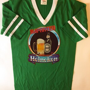 Pu&ograve; includere: Maglietta verde a maniche corte con strisce bianche sulle maniche. La maglietta ha un collo a V e presenta una grafica di una bottiglia di birra e un bicchiere con il testo "IMPORTED" e "Heineken" in rosso e oro.
