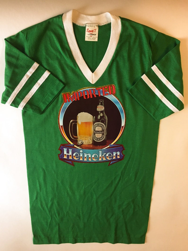 Pu&ograve; includere: Maglietta verde a maniche corte con strisce bianche sulle maniche. La maglietta ha un collo a V e presenta una grafica di una bottiglia di birra e un bicchiere con il testo "IMPORTED" e "Heineken" in rosso e oro.