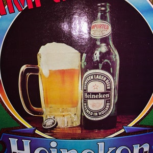 Pu&ograve; includere: Una maglietta verde con una grande immagine colorata di una bottiglia di birra Heineken e un bicchiere di birra. Il testo "IMPORTED" &egrave; stampato in rosso sopra l'immagine. Il testo "Heineken" &egrave; stampato su uno striscione blu sotto l'immagine.