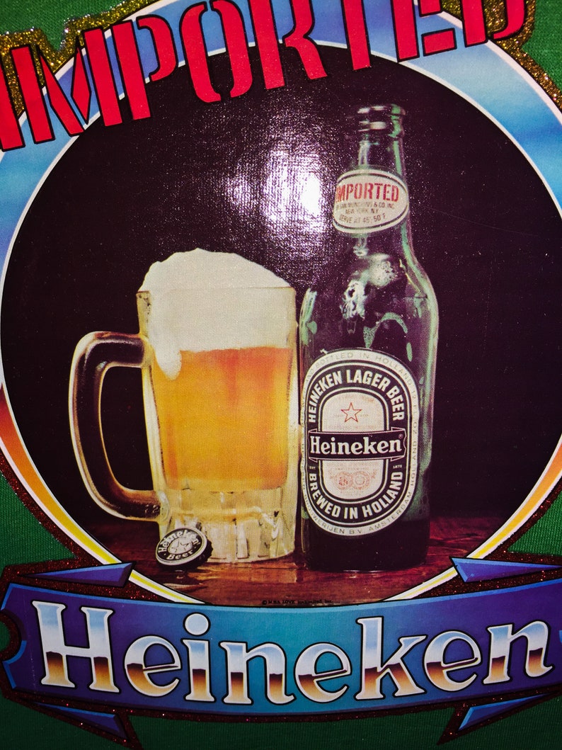 Pu&ograve; includere: Una maglietta verde con una grande immagine colorata di una bottiglia di birra Heineken e un bicchiere di birra. Il testo "IMPORTED" &egrave; stampato in rosso sopra l'immagine. Il testo "Heineken" &egrave; stampato su uno striscione blu sotto l'immagine.