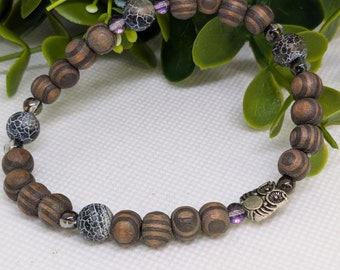 May include: A wooden beaded bracelet with brown and black striped beads, grey agate beads, and a silver owl charm. The bracelet is on a white surface.