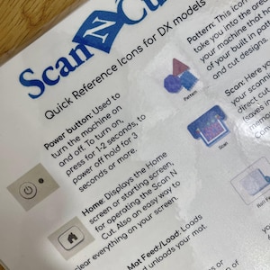 May include: A white instruction sheet with blue text and icons for a ScanNCut machine. The sheet explains the functions of the power button, home screen, scan button, mat feed, and pattern button. The text reads "Quick Reference Icons for DX models".