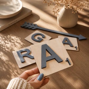 May include: Wooden Scrabble tiles with gray letters spelling "RAGA" and a gray arrow. The letter tiles are on a wooden surface. A hand is holding one of the tiles. A vase with dried flowers is in the background.