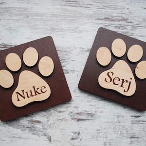 Puede incluir: Dos letreros cuadrados de madera marr&oacute;n con un dise&ntilde;o de huella de pata. La huella de pata est&aacute; hecha de madera clara y tiene los nombres "Nuke" y "Serj" grabados.