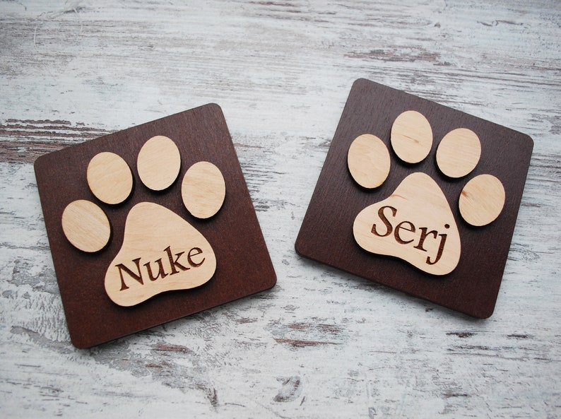 Puede incluir: Dos letreros cuadrados de madera marr&oacute;n con un dise&ntilde;o de huella de pata. La huella de pata est&aacute; hecha de madera clara y tiene los nombres "Nuke" y "Serj" grabados.