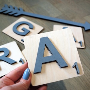 May include: Wooden Scrabble tiles with gray letters and numbers. An arrow-shaped decoration is also present. The letter tiles include the letters A, G, and R, each with a value of 1. The arrow and letters are in a gray color.