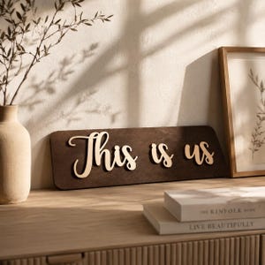 May include: A rectangular dark brown wooden sign featuring the light-coloured, raised script text "This is us" centred on the front surface. The sign has rounded corners and a smooth finish, displaying a clear contrast between the dark base and lettering.