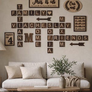 May include: Wooden wall decor featuring scrabble-style tiles spelling out "FAMILY", "FRIENDS", and names, along with dates and phrases like "This is us" and "Our nest". The decor is arranged above a beige sofa and wooden coffee table.