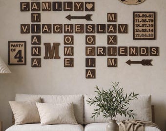 May include: Wooden wall decor featuring scrabble-style tiles spelling out "FAMILY", "FRIENDS", and names, along with dates and phrases like "This is us" and "Our nest". The decor is arranged above a beige sofa and wooden coffee table.
