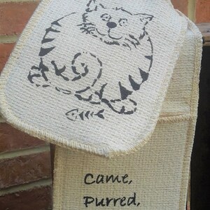May include: Off-white oven mitt with a cat illustration and the words "Came, Purred, Conquered." The cat is drawn in black ink, with a fish skeleton below. The mitt is hanging against a brick wall.