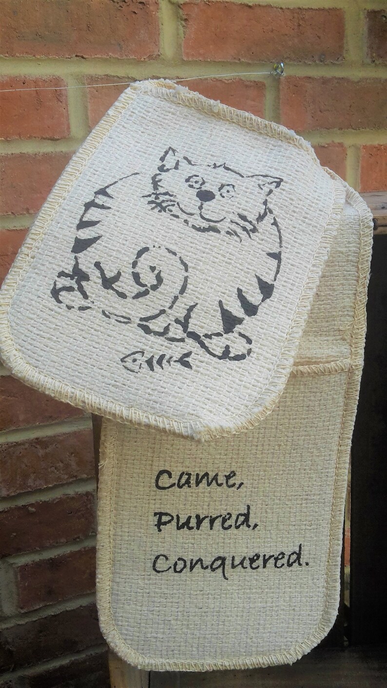 May include: Off-white oven mitt with a cat illustration and the words "Came, Purred, Conquered." The cat is drawn in black ink, with a fish skeleton below. The mitt is hanging against a brick wall.