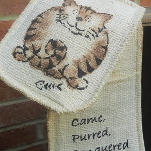 May include: A beige double oven mitt with a cartoon cat illustration in brown and black. The text "Came, Purred, Conquered" is printed in black on the lower mitt.