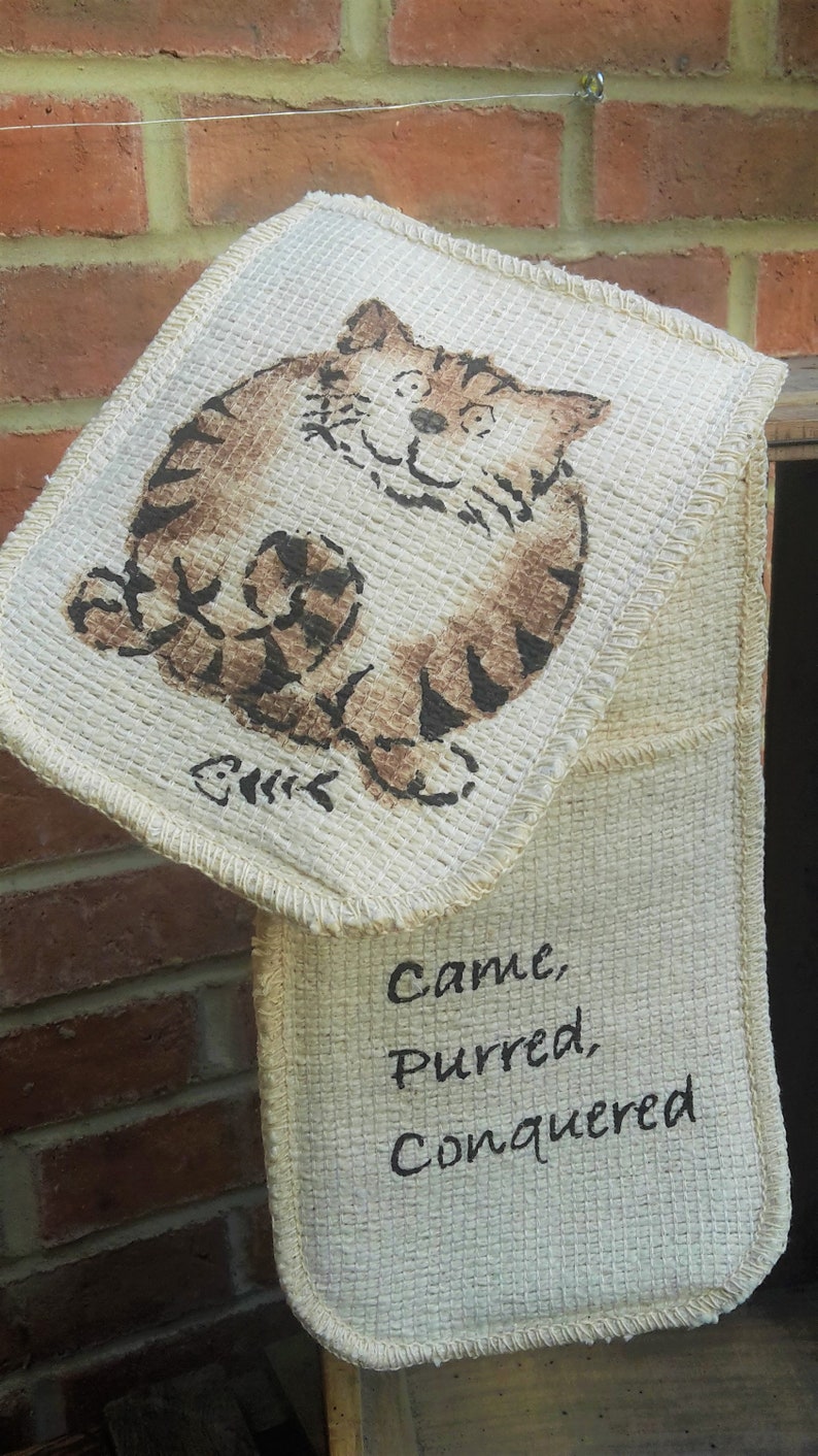 May include: A beige double oven mitt with a cartoon cat illustration in brown and black. The text "Came, Purred, Conquered" is printed in black on the lower mitt.