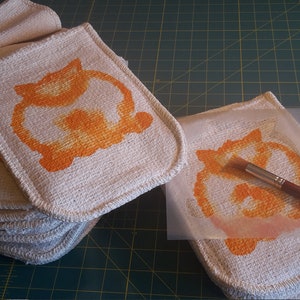 May include: A stack of off-white, square potholders with rounded corners, each featuring an orange cat design. A paintbrush and a stencil with the same design are also visible, suggesting a handmade craft project.