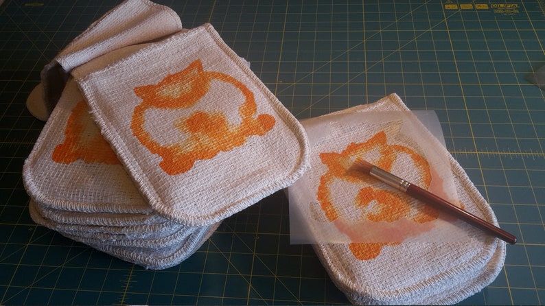 May include: A stack of off-white, square potholders with rounded corners, each featuring an orange cat design. A paintbrush and a stencil with the same design are also visible, suggesting a handmade craft project.