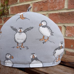 May include: A grey tea cosy with a white trim and a pattern of puffins on a grey background.