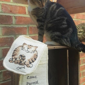 May include: A tabby cat sits atop a wooden crate, with a matching oven mitt and pot holder set featuring a cartoon cat design. The text "Came, Purred, Conquered" is printed on the oven mitt. The background includes a brick wall and greenery.