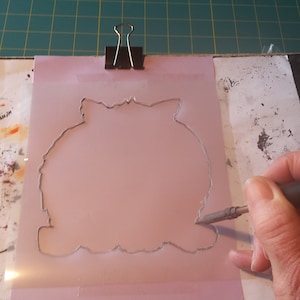 May include: A pink stencil with a detailed outline is held in place with a black binder clip. A hand uses a craft knife to cut along the stencil's edge. The cutting surface is a green grid.