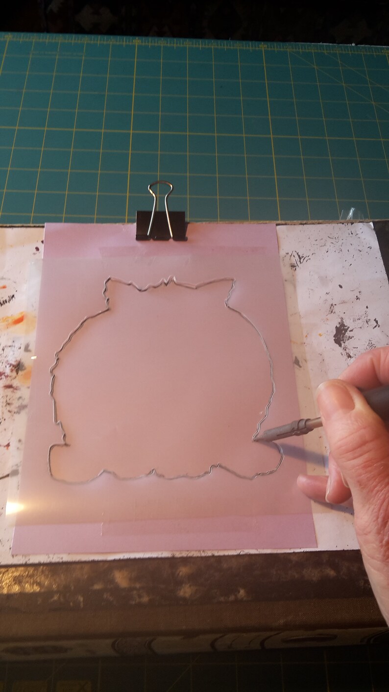 May include: A pink stencil with a detailed outline is held in place with a black binder clip. A hand uses a craft knife to cut along the stencil's edge. The cutting surface is a green grid.