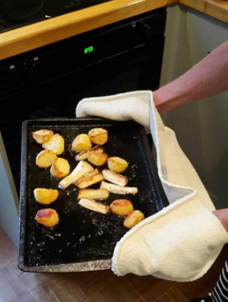 May include: A black baking sheet holds roasted potatoes and parsnips. The vegetables are golden brown and appear freshly cooked. The sheet is held with white oven mitts, suggesting a hot dish just out of the oven.
