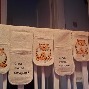 May include: Beige oven mitts with a cartoon orange tabby cat design. Each mitt has the words "Came, Purred, Conquered." printed on it. The mitts are hanging on a white railing.