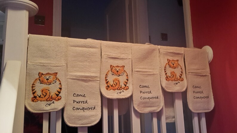 May include: Beige oven mitts with a cartoon orange tabby cat design. Each mitt has the words "Came, Purred, Conquered." printed on it. The mitts are hanging on a white railing.
