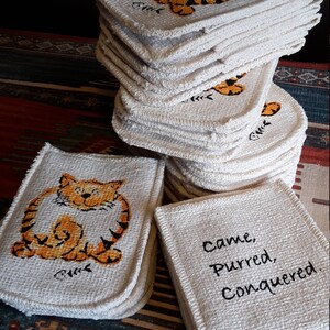 May include: A stack of square, off-white coasters with a woven texture. Each coaster features a cartoon tiger in orange and black. One stack has the text "Came, Purred, Conquered."