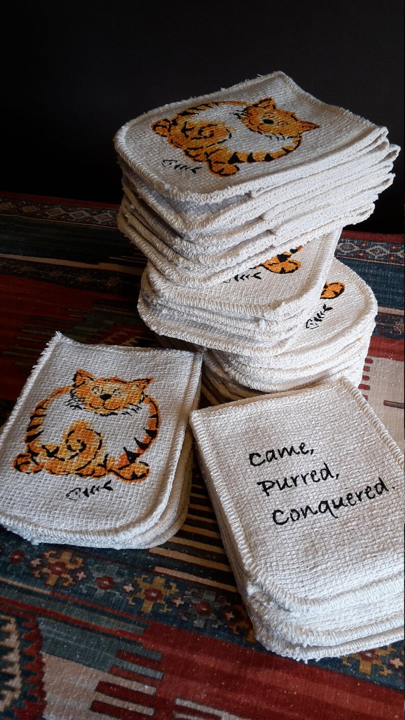 May include: A stack of square, off-white coasters with a woven texture. Each coaster features a cartoon tiger in orange and black. One stack has the text "Came, Purred, Conquered."