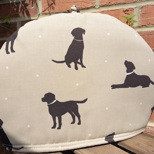 May include: A beige tea cosy with a repeating pattern of black Labrador dog silhouettes and small white dots. The cosy has a loop at the top for hanging. The fabric design features dogs in different poses.