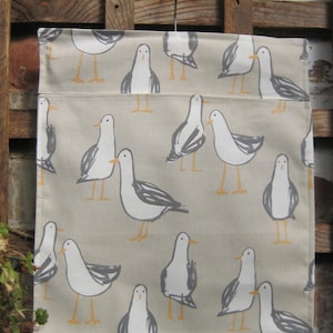 May include: A beige fabric tote bag with a repeating pattern of white seagulls. The bag has a pocket on the front and a metal hook at the top.