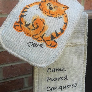 May include: A beige oven mitt with a cartoon orange cat design and the words "Came, Purred, Conquered." The cat illustration includes a fishbone symbol. The mitt is hanging against a brick wall.