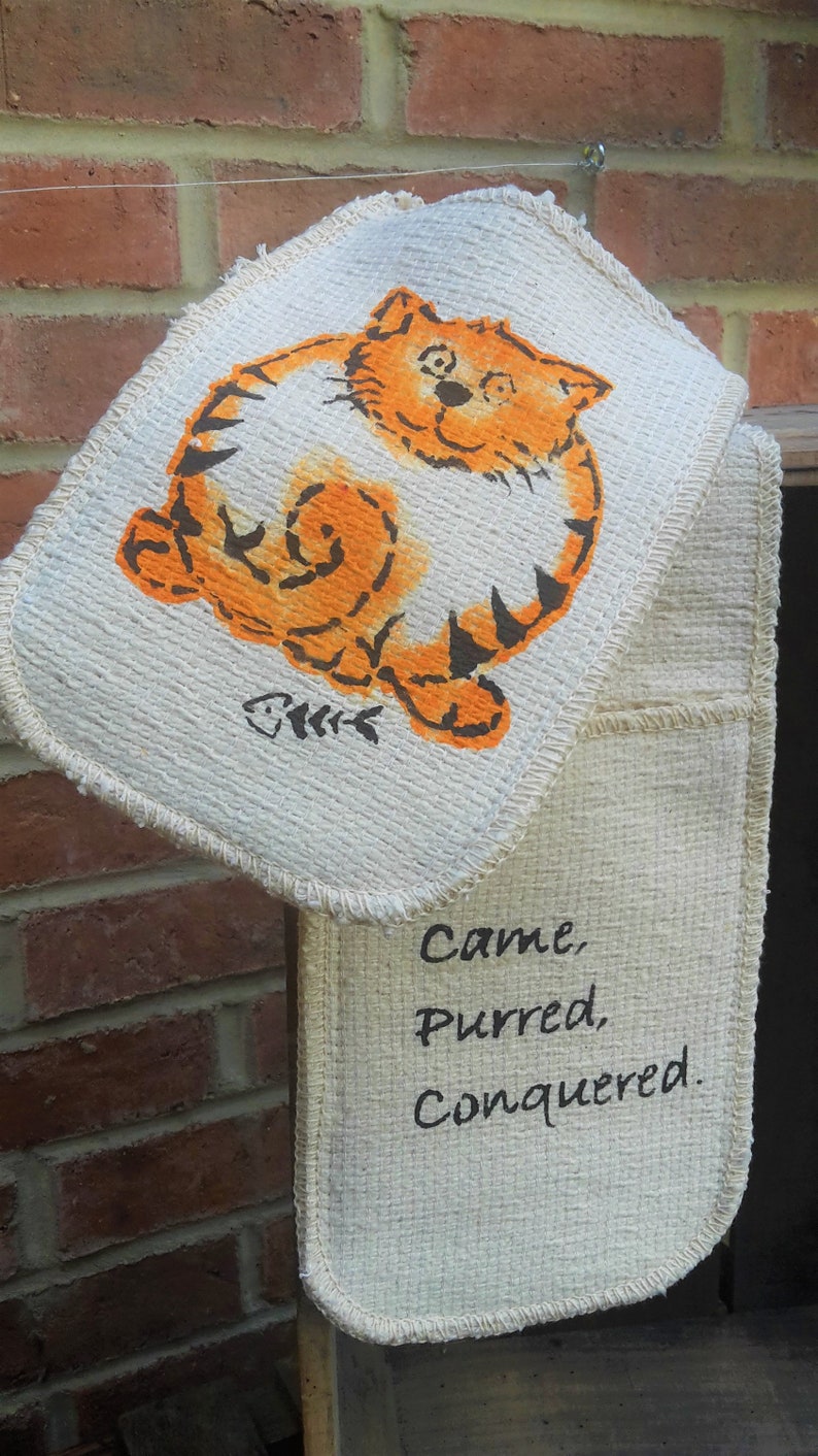 May include: A beige oven mitt with a cartoon orange cat design and the words "Came, Purred, Conquered." The cat illustration includes a fishbone symbol. The mitt is hanging against a brick wall.