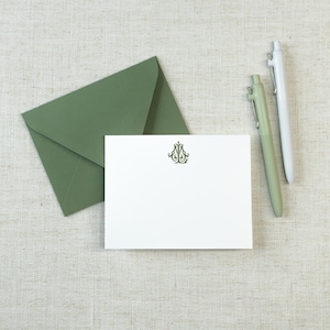 May include: A white rectangular note card featuring a centered dark green ornate monogram, accompanied by a matching sage green envelope and two pens, one in sage green and one in white, arranged on a neutral textured fabric surface.