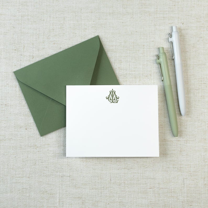May include: A white rectangular note card featuring a centered dark green ornate monogram, accompanied by a matching sage green envelope and two pens, one in sage green and one in white, arranged on a neutral textured fabric surface.