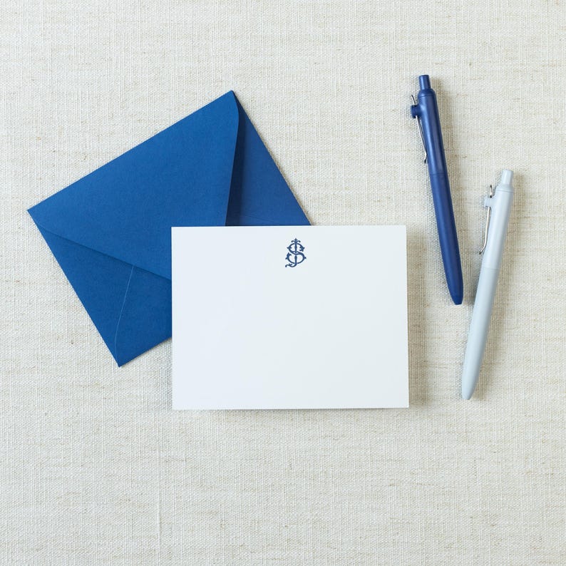 May include: A white rectangular note card featuring a dark blue monogram of the letters S and J centered at the top, accompanied by a matching dark blue envelope, a dark blue retractable ballpoint pen, and a light gray retractable ballpoint pen.