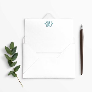 May include: A white envelope with a white card inside. The card has a teal monogram of the letter "M" in a decorative font. A brown pen is lying on a white surface next to the envelope.