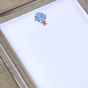 May include: White stationery paper with a blue hydrangea floral design and a red bow. The paper is set inside a clear acrylic frame, resting on a neutral-toned fabric surface.