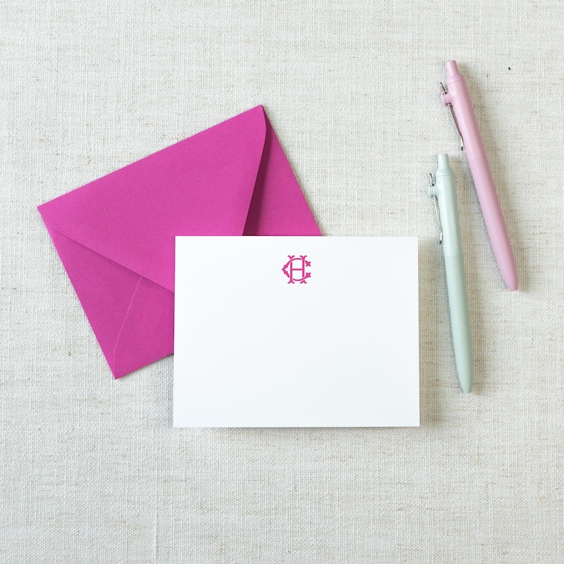 May include: A white rectangular note card featuring a centered magenta monogram with the letters C and H, accompanied by a matching magenta envelope and two pens, one in light pink and one in light blue, arranged on a textured fabric surface.