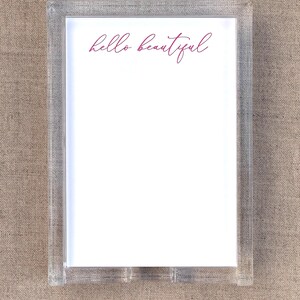 May include: Clear acrylic frame with a white insert. The words "hello beautiful" are written in a cursive, magenta font across the top of the insert. The frame is rectangular and sits on a neutral, textured surface.