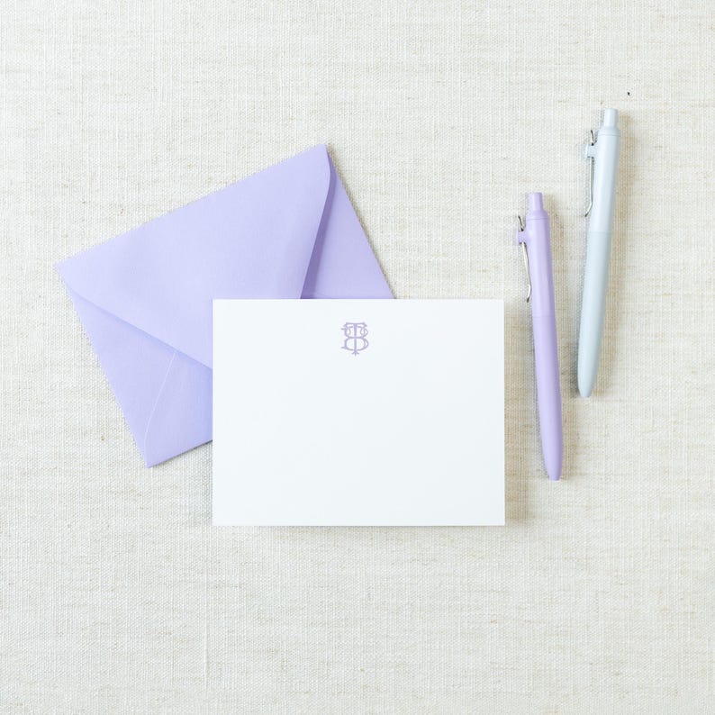 May include: A white rectangular note card featuring a centered purple monogram of the letters SB, accompanied by a matching light purple envelope and two retractable pens in light purple and light blue with silver clips and accents.
