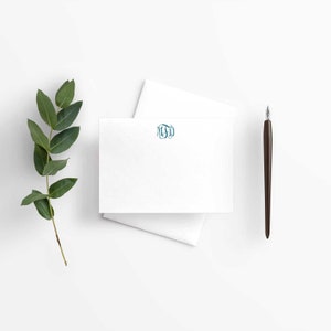 May include: A white card with a teal blue monogram "MSD" printed on it. The card is sitting on a white surface with a green leafy branch and a brown pen.