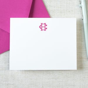 May include: A white rectangular note card featuring a centered hot pink monogram with the letters C and H intertwined. The card is positioned next to a bright pink envelope and a light-colored pen, creating a clean and simple stationery set.