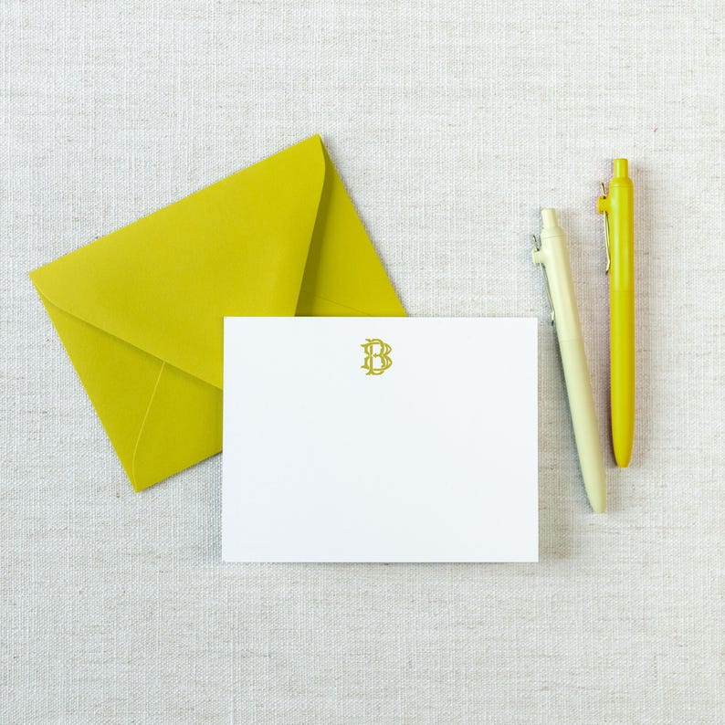 May include: A white rectangular note card featuring a centered olive green letter B monogram, accompanied by a matching olive green envelope and two pens, one in a pale yellow and the other in a vibrant yellow, arranged on a textured surface.