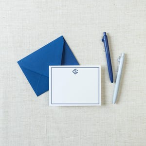 May include: A white rectangular note card with a dark blue border and a central monogram, accompanied by a matching dark blue envelope, a dark blue retractable ballpoint pen, and a light gray retractable ballpoint pen.